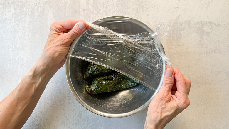 Covering grilled poblano peppers in stainless steel bowl with plastic wrap